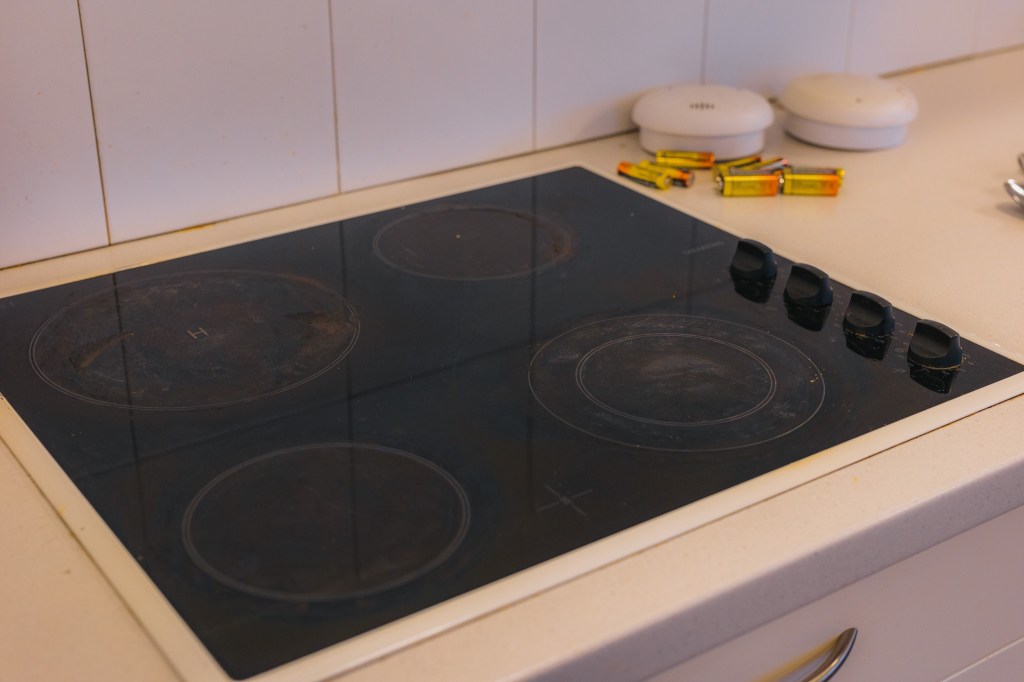 A black electric stovetop with four burners is set into a white countertop. Nearby are two smoke detectors and several AA batteries.