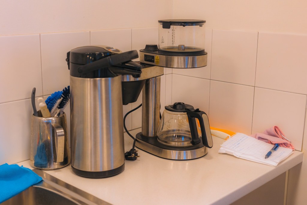 A stainless steel coffee maker and thermos sit on a white kitchen counter. Nearby are cleaning brushes, a notebook, pen, and a folded cloth.