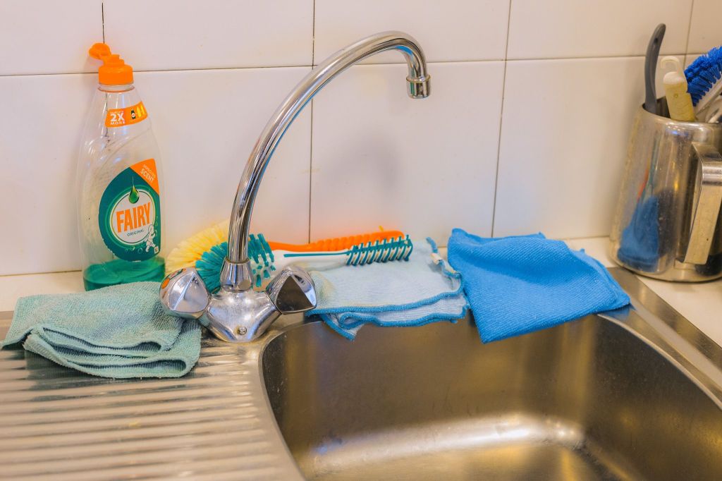 Kitchen sink with a chrome faucet surrounded by cleaning items: sponges, brushes, and a dish soap bottle.