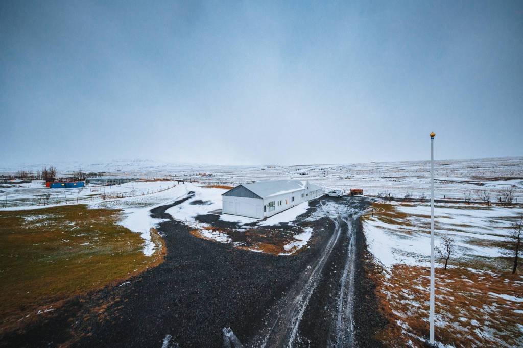 Remote snowy landscape with a long white building centered, surrounded by partially snow-covered ground and distant hills under an overcast sky.