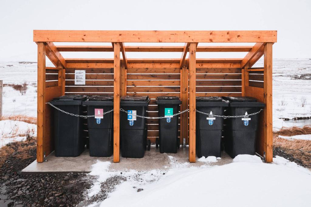 Five chained black recycling bins under a wooden shelter on snow. Each bin is labeled for sorting.
