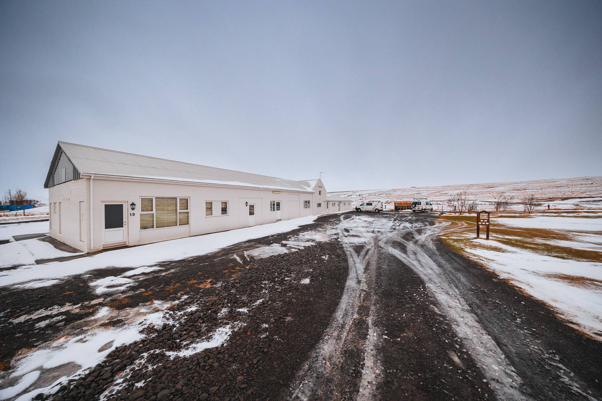 Long white building on a snowy, overcast day with dark tire tracks leading to parked vehicles.