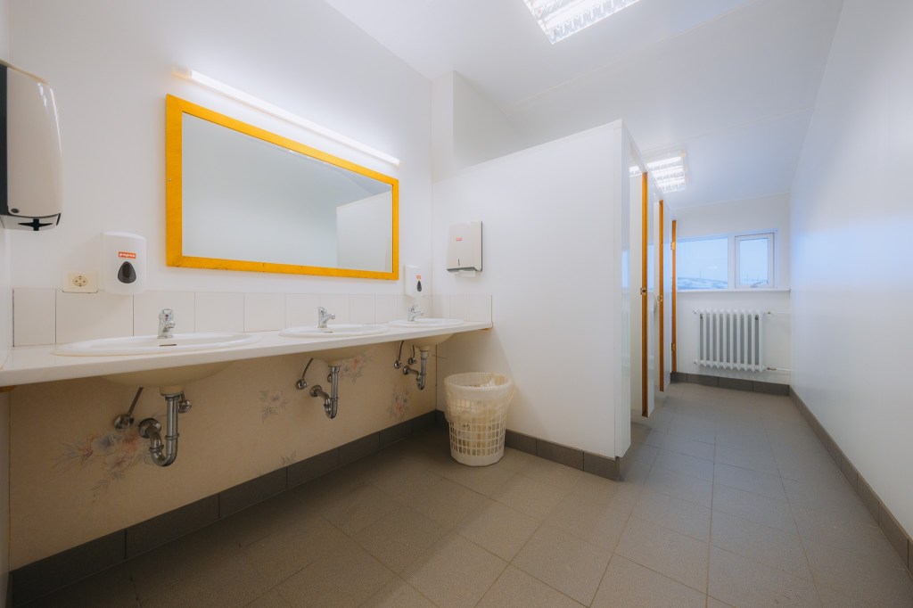 A clean, spacious public restroom with white walls and floor tiles. Three sinks are beneath a large mirror with a yellow frame.