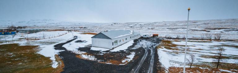 Remote snowy landscape with a long white building centered, surrounded by partially snow-covered ground and distant hills under an overcast sky.