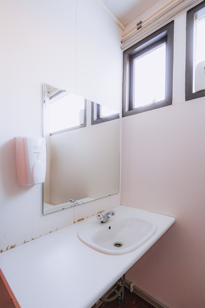 Small bathroom with a white sink and mirror. Two windows provide natural light. A wall-mounted soap dispenser is next to the mirror.