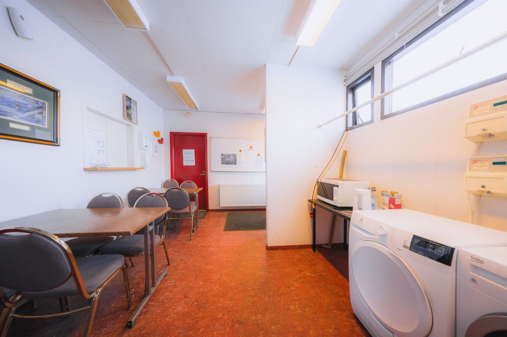 Laundry room with a bright, clean ambiance. Features a washer, dryer, small table, chairs, and a red door. Wall with notices and small appliances.