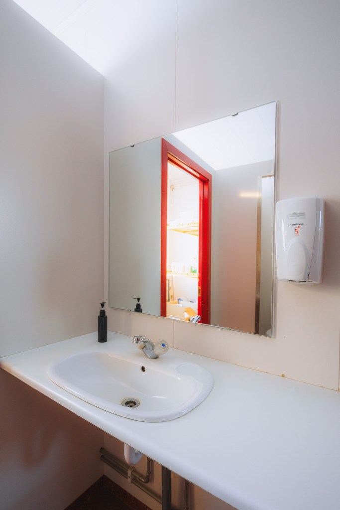 A bathroom scene with a white sink and a chrome faucet on a counter. Above, a large mirror reflects a doorway with a bright red frame. A white soap dispenser is mounted on the wall beside the mirror, and a black soap bottle is on the counter