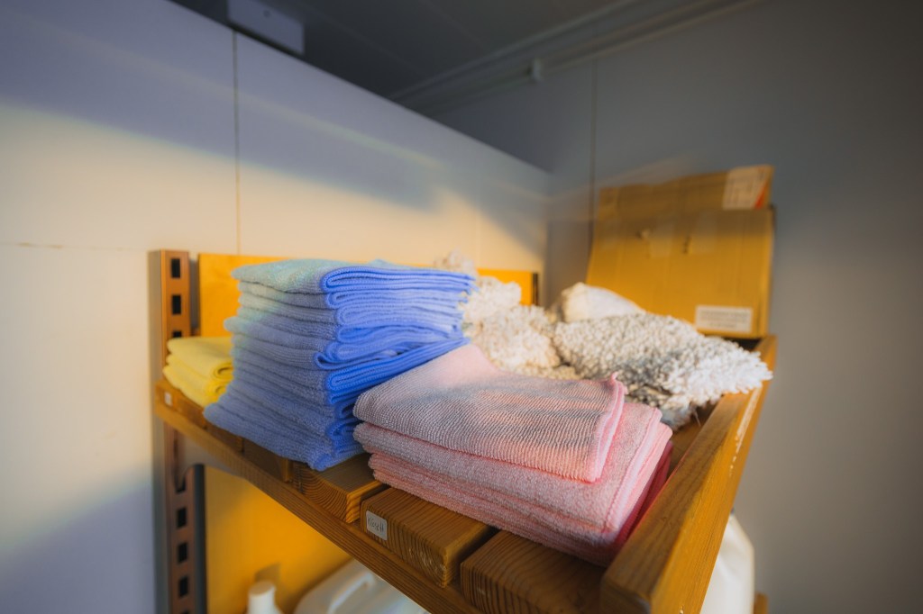 Stacks of folded microfiber cloths in blue, pink, and yellow rest on a wooden shelf, bathed in warm sunlight.