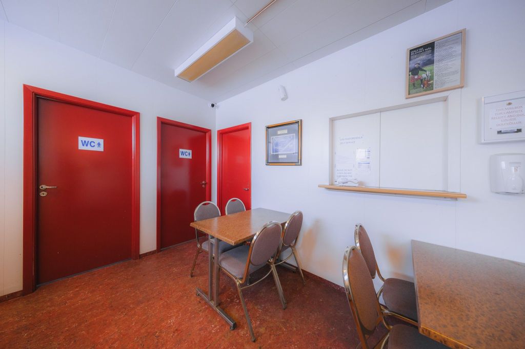 A room with red doors marked "WC," a brown table with chairs, white walls, and various framed notices.
