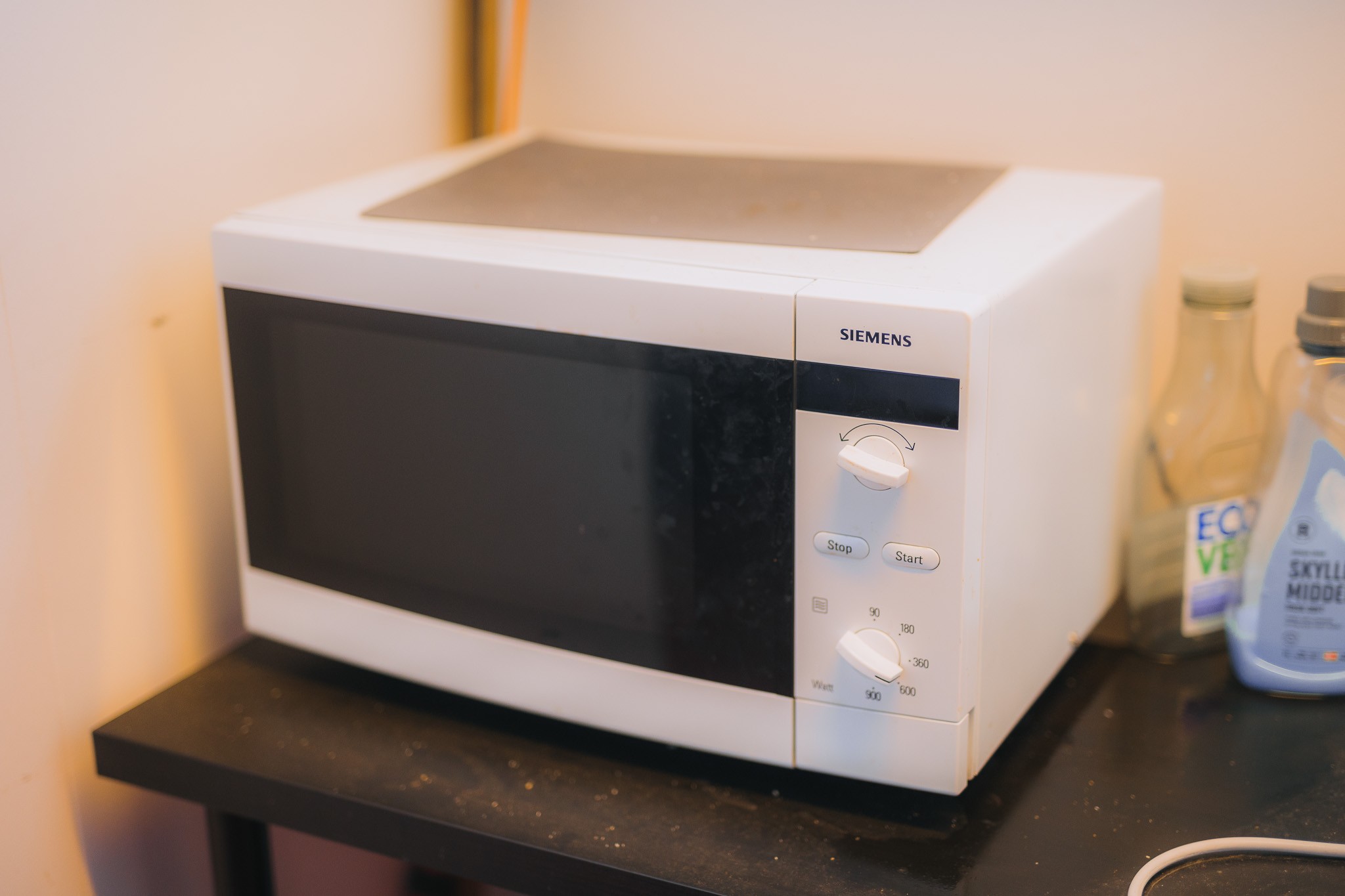 A white microwave oven with a black door and control knobs is on a black countertop. Nearby are two bottles, one labeled "Eco," in a well-lit kitchen corner.