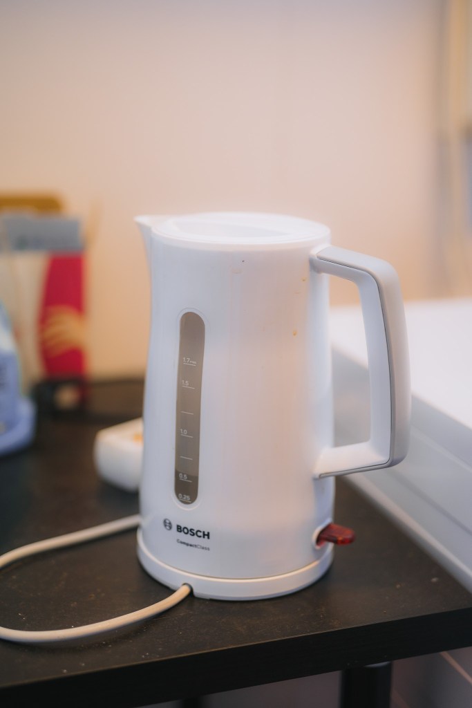 White electric kettle with a measurement window, marked Bosch, sits on a dark countertop in a kitchen setting. Nearby items are slightly blurred.