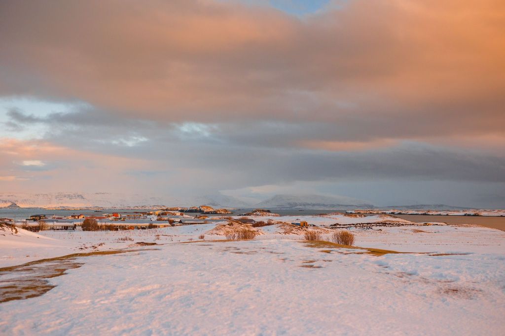 Snow-covered landscape with distant icy mountains under a cloudy, orange-hued sky during sunset.