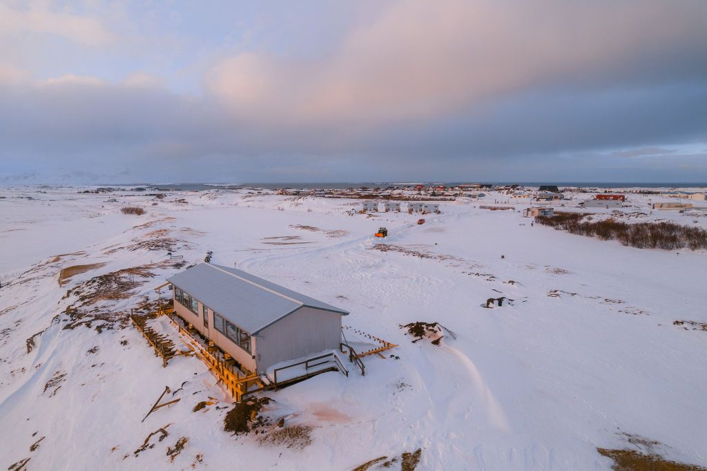 A solitary gray building stands on a snowy landscape under a cloudy sky, with a distant cluster of buildings.