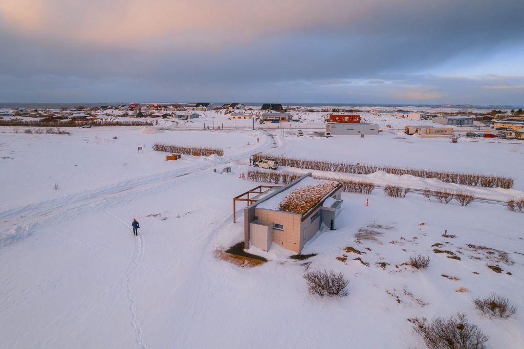 Snowy landscape with scattered houses under a pastel sky. A lone person walks through the snow near a small building.