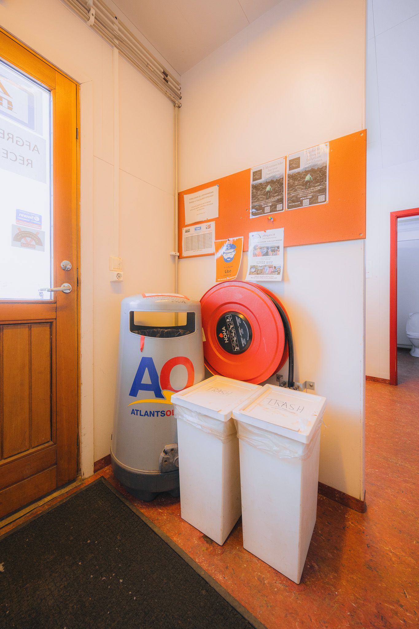 A hallway corner features a wooden door, a waste bin with a logo, white recycling bins, a red hose reel, and an orange bulletin board with notices.