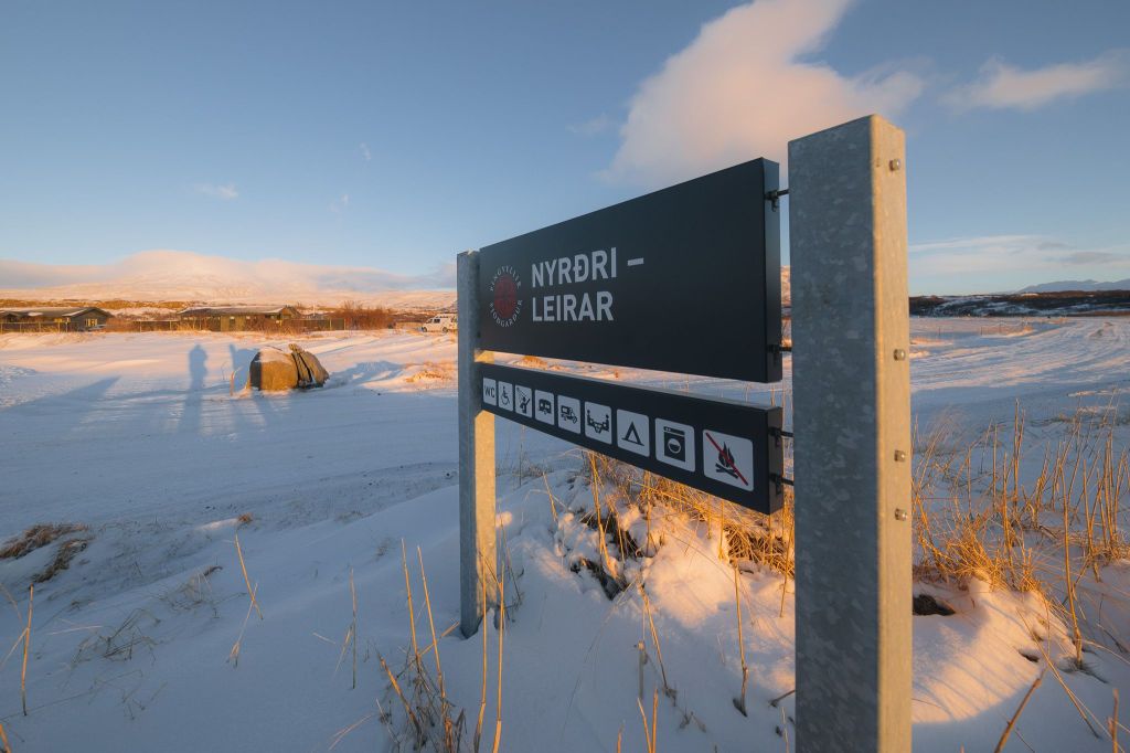 Snowy landscape with a sign reading "NYRÐRI LEIRAR" displaying various icons. Long shadows cast by the setting sun create a serene, wintry ambiance.