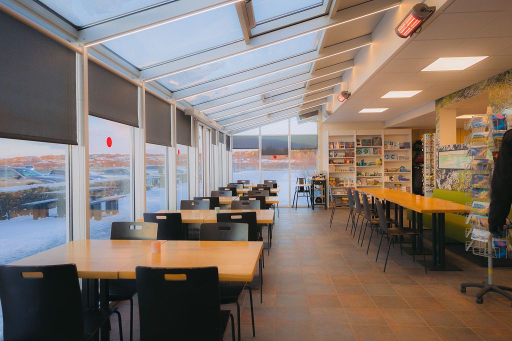 Sunlit cafeteria interior with large windows showing a snowy landscape. Wooden tables and black chairs are neatly arranged.