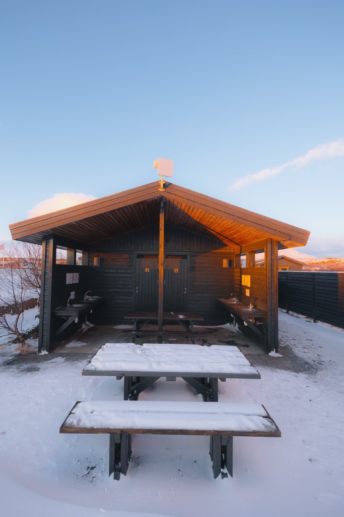 A wooden shelter with picnic tables sits under a snowy, clear blue sky.