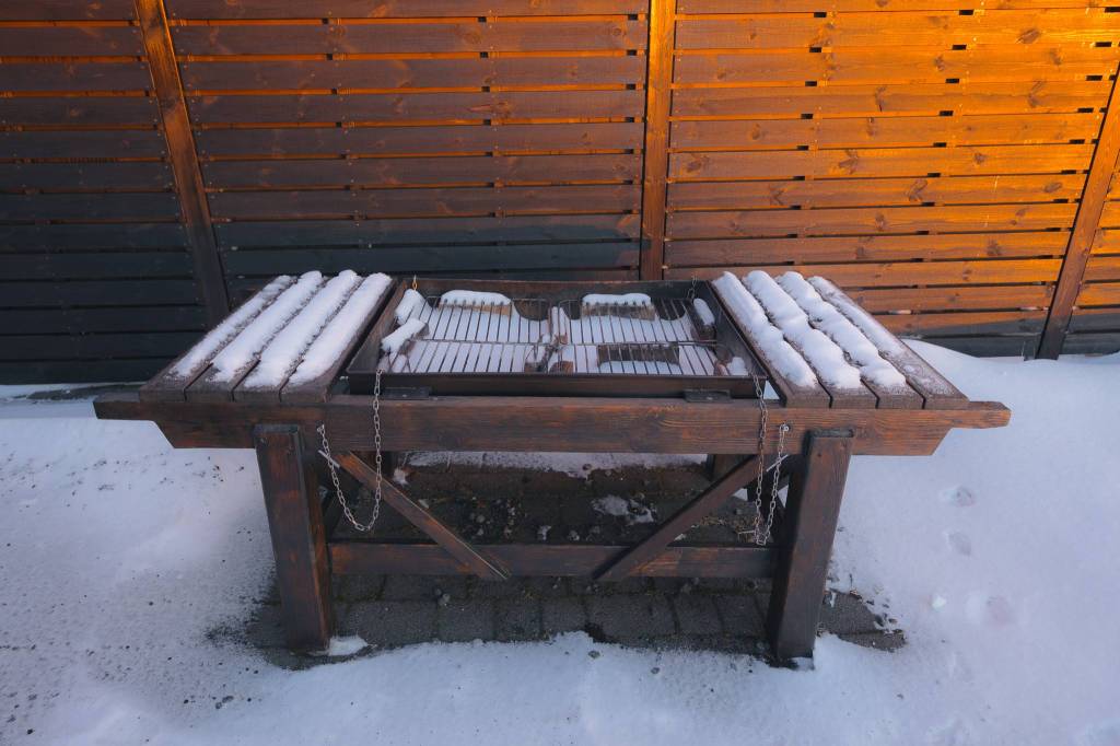 A snow-covered wooden grill table stands against a wooden fence, with a wintery atmosphere.