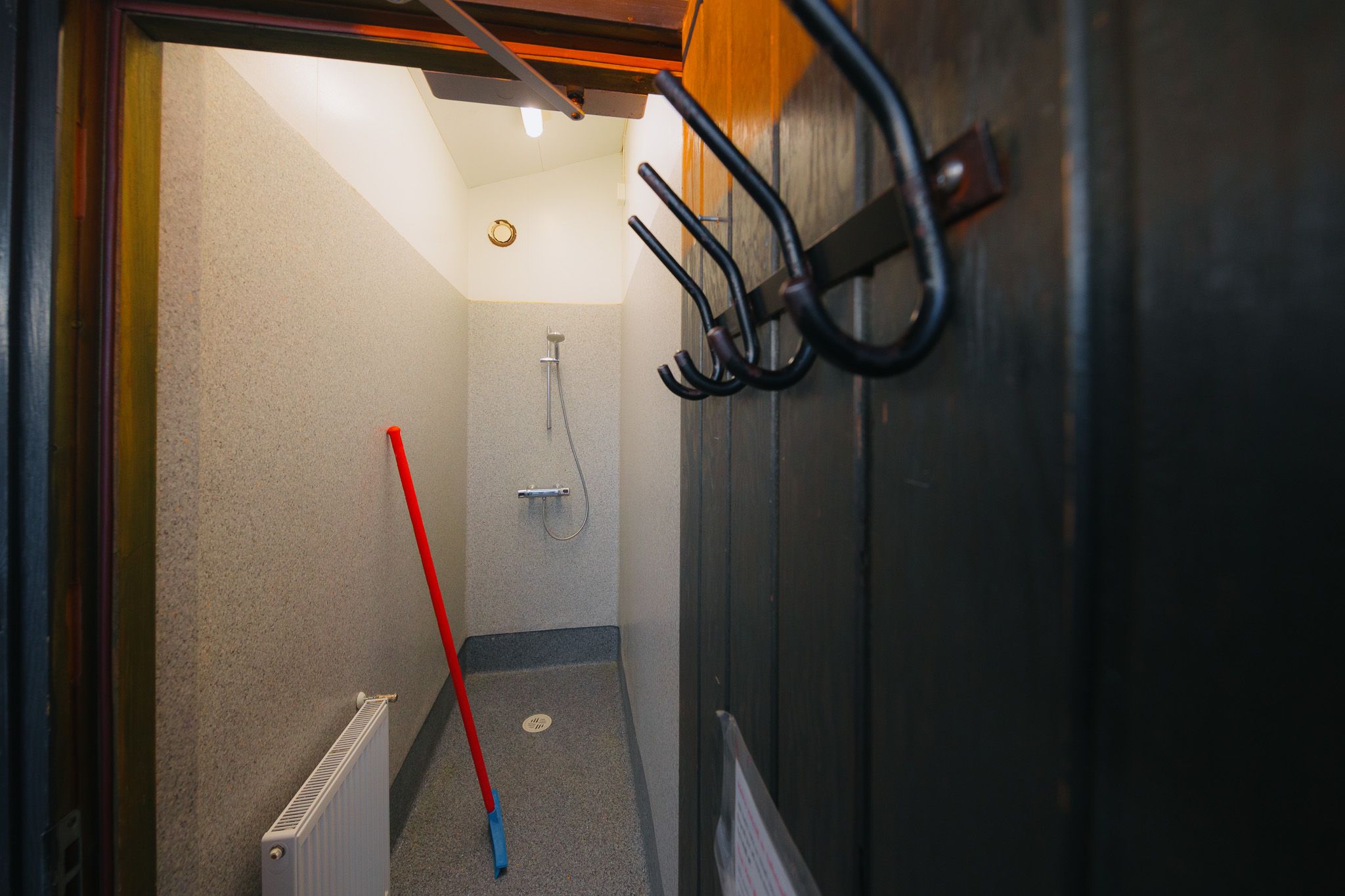 Small, dimly lit shower room with a wall-mounted showerhead. A red mop leans against the wall. Black coat hooks are visible in the foreground.