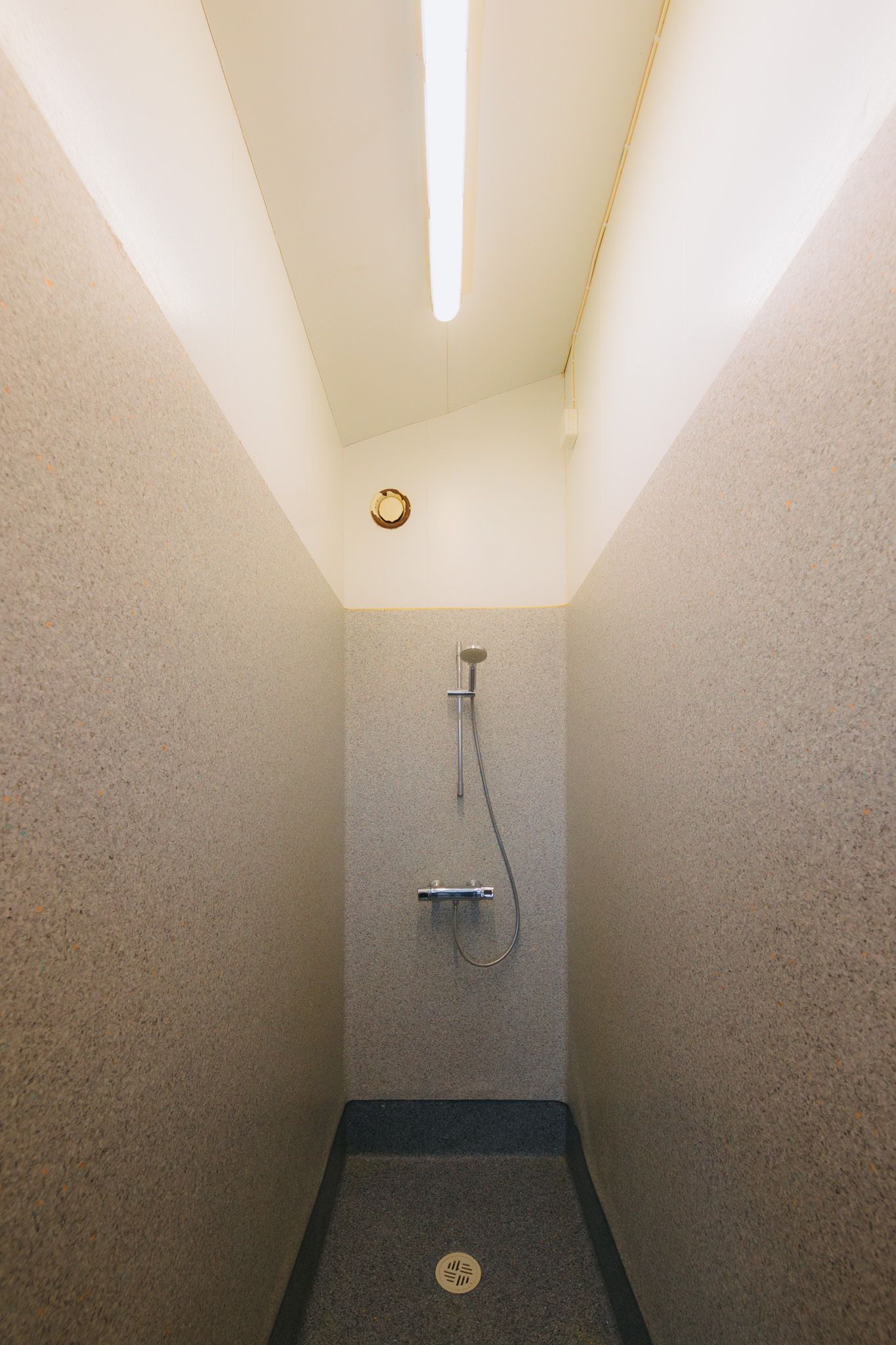 Small, minimalist shower with beige tile walls and a ceiling light. Features a wall-mounted showerhead with a hose and a drain on the floor.