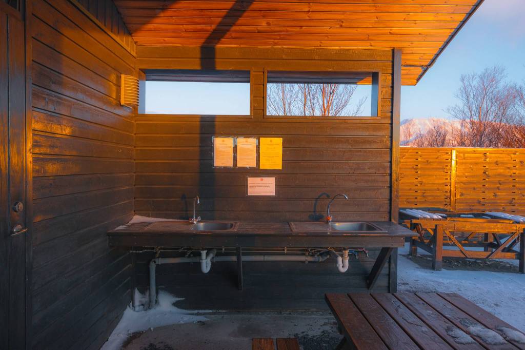 Outdoor kitchen with two metal sinks and wood surfaces, surrounded by wood paneling. Evening sunlight casts warm tones, with snowy mountains in the background.