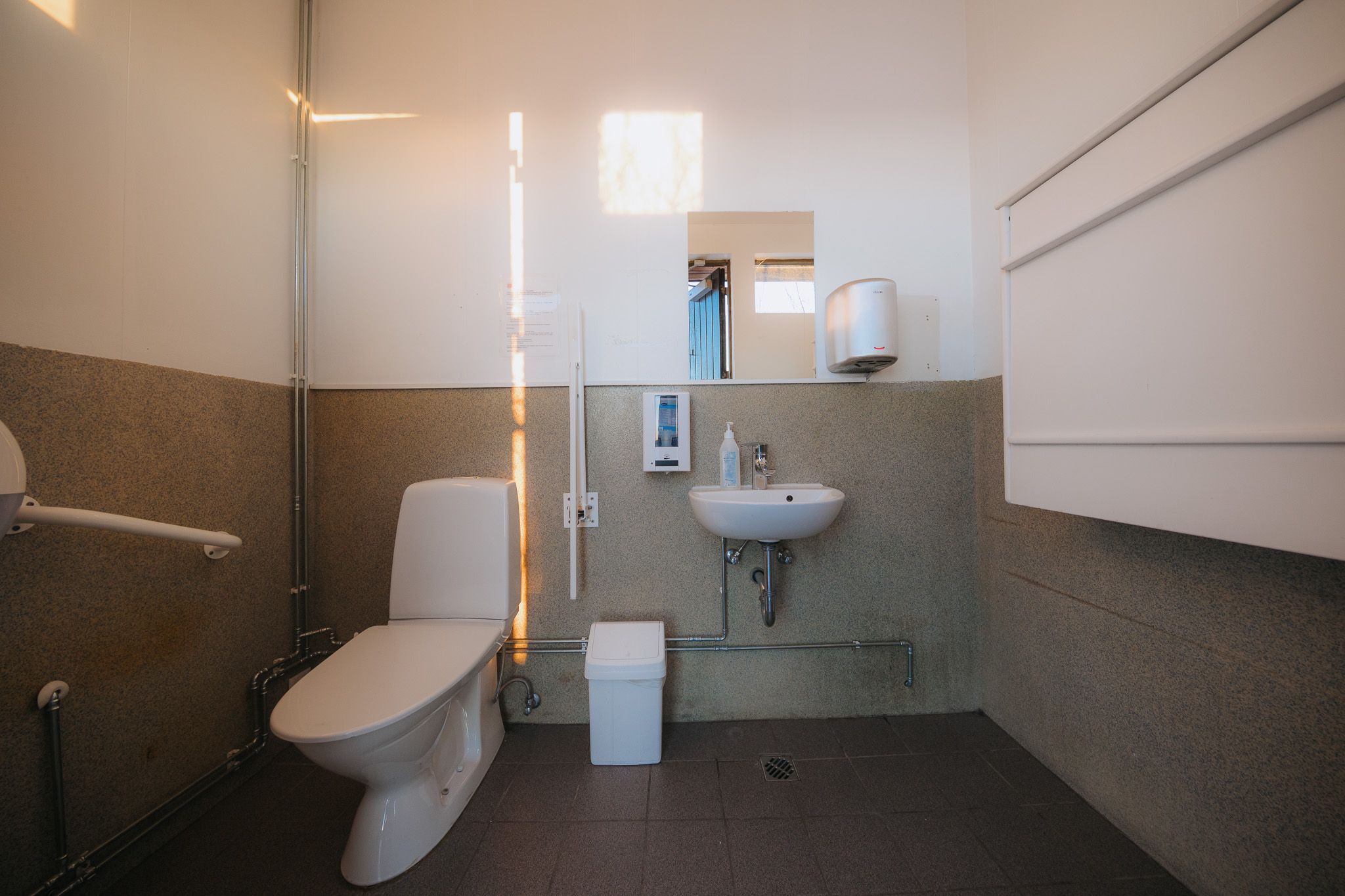A clean, minimalist bathroom with a toilet, small sink, mirror, and dispenser.
