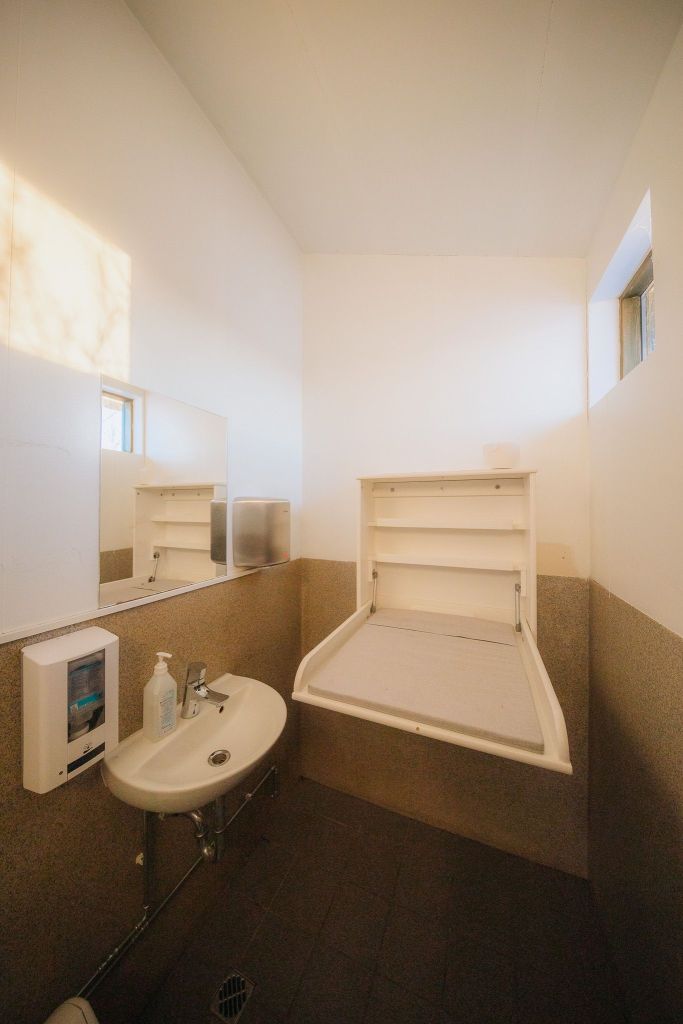 A small, brightly lit bathroom with white walls and a gray tiled lower half. It features a sink with a soap dispenser and a mirror above. A changing table is mounted on the right wall, unfolded and ready for use.