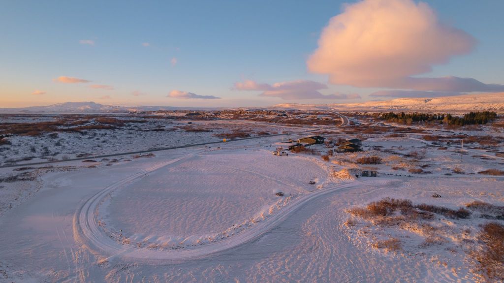 A snow-covered landscape at sunrise, with a winding road leading to a few scattered buildings.