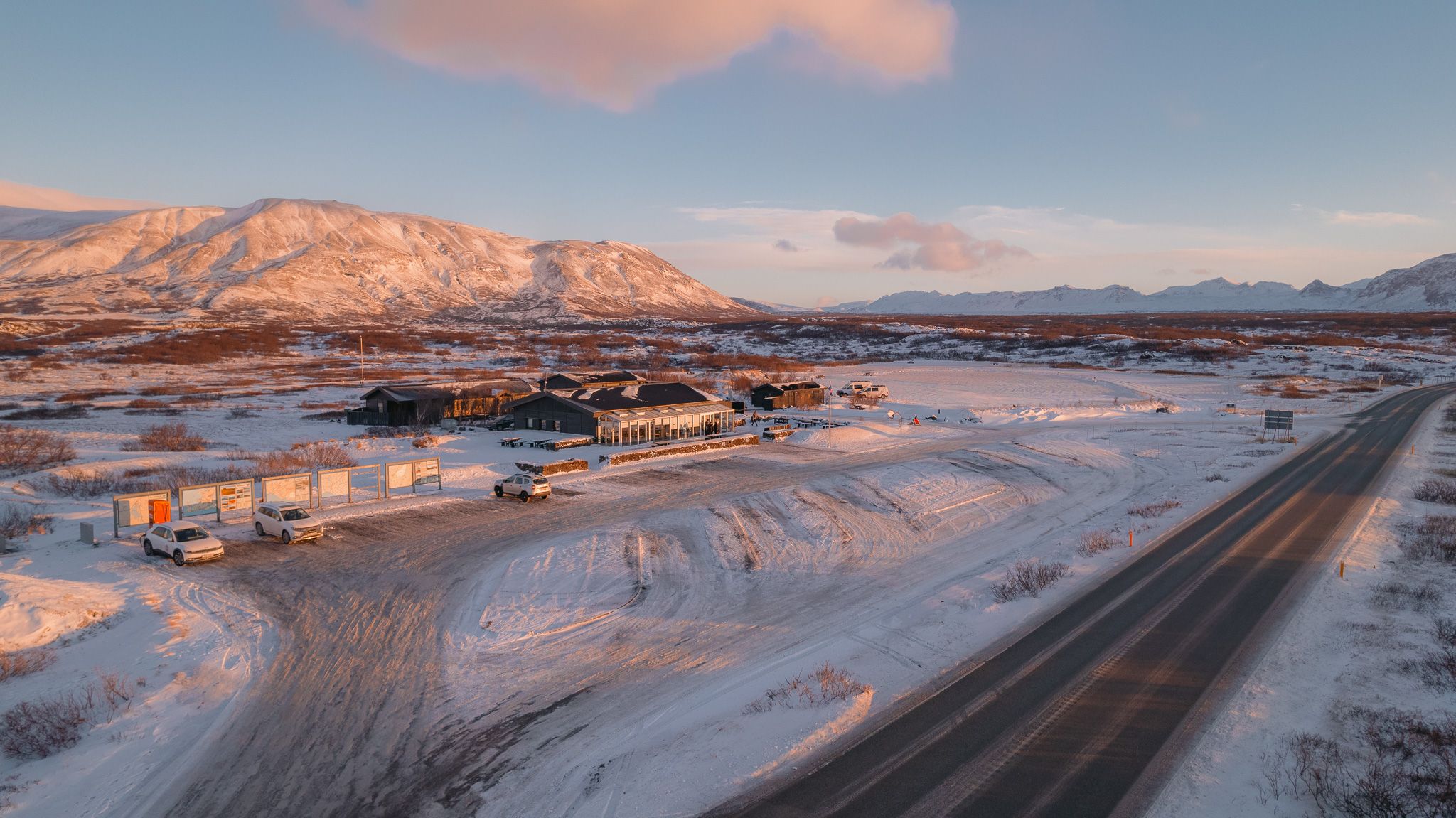 A snowy landscape at dawn featuring a remote highway and buildings. Rugged mountains in the background under a pastel sky.