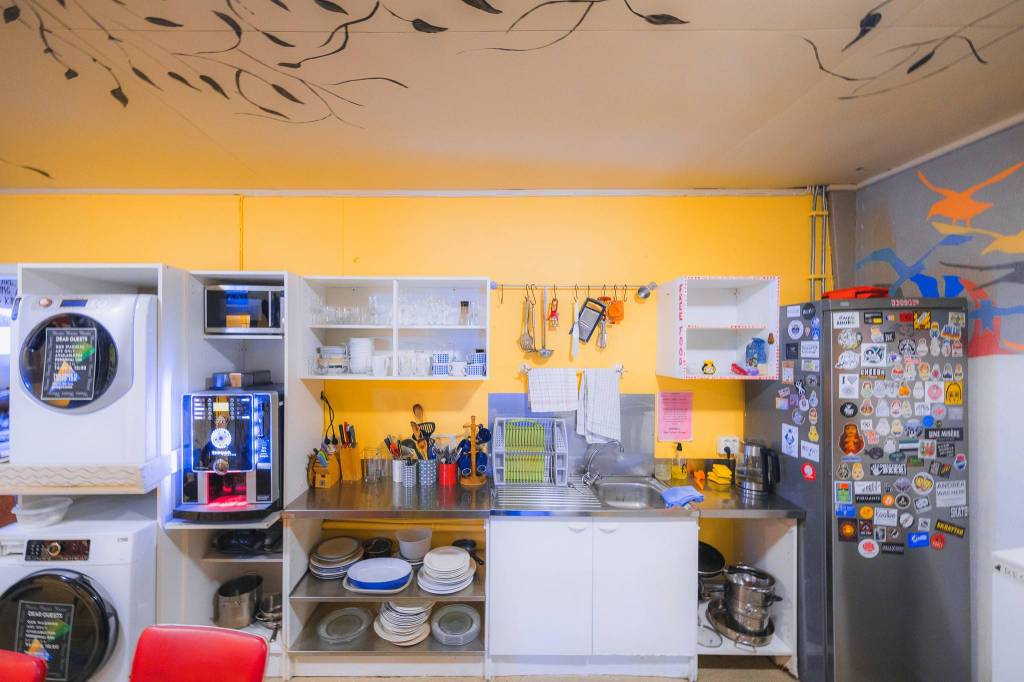 A cozy, colorful kitchen with yellow walls features open shelves holding plates and glasses, a fridge adorned with stickers, and a vibrant mural on the ceiling.