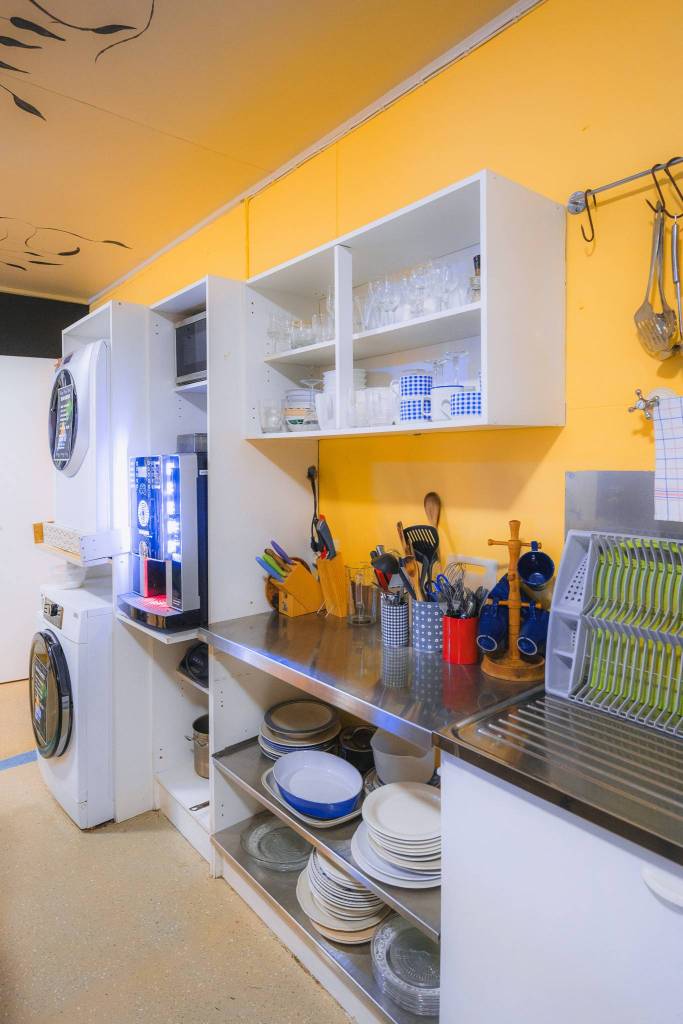 A brightly lit kitchen with orange walls features a coffee machine, stacked dishes, and utensils on a stainless steel counter.
