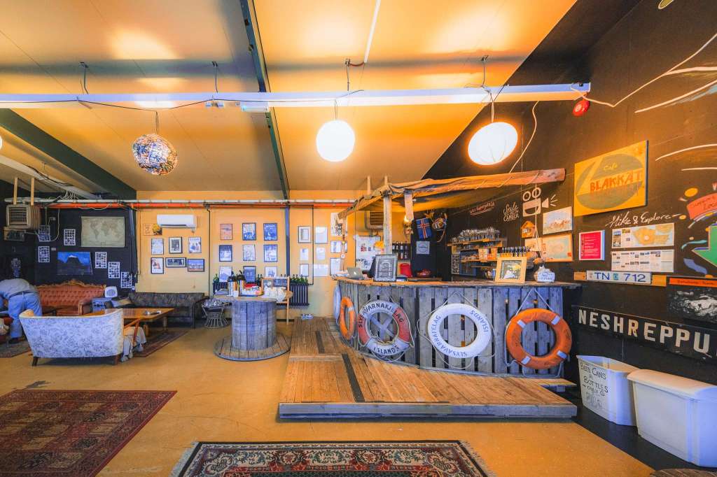 A cozy interior of a café with warm lighting, wooden decor, and nautical-themed elements. Walls feature framed photos and a chalkboard menu. Comfortable seating and rugs add a welcoming atmosphere.