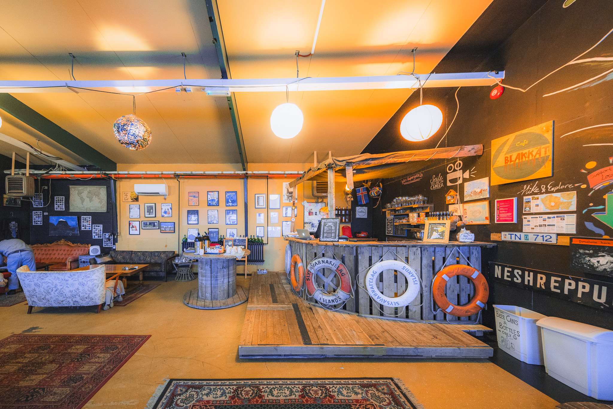 A cozy interior of a café with warm lighting, wooden decor, and nautical-themed elements. Walls feature framed photos and a chalkboard menu. Comfortable seating and rugs add a welcoming atmosphere.