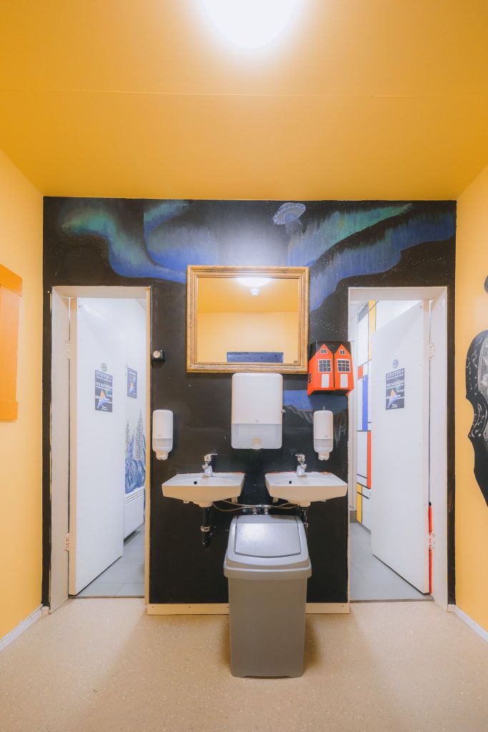 Restroom with vibrant yellow walls and two open stalls. Central black wall features a framed mirror, two sinks, soap dispensers, and a red thing.