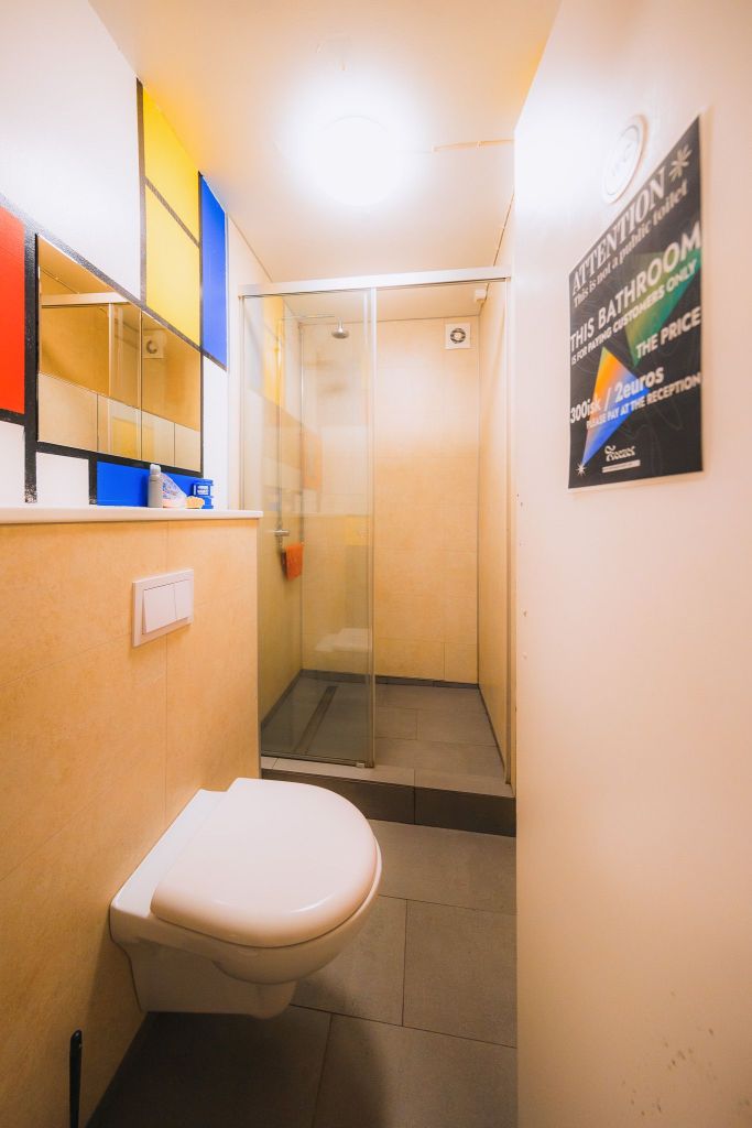 A small, well-lit bathroom with beige walls features a floating toilet, glass shower, colorful art panels, and a sign about bathroom charges.