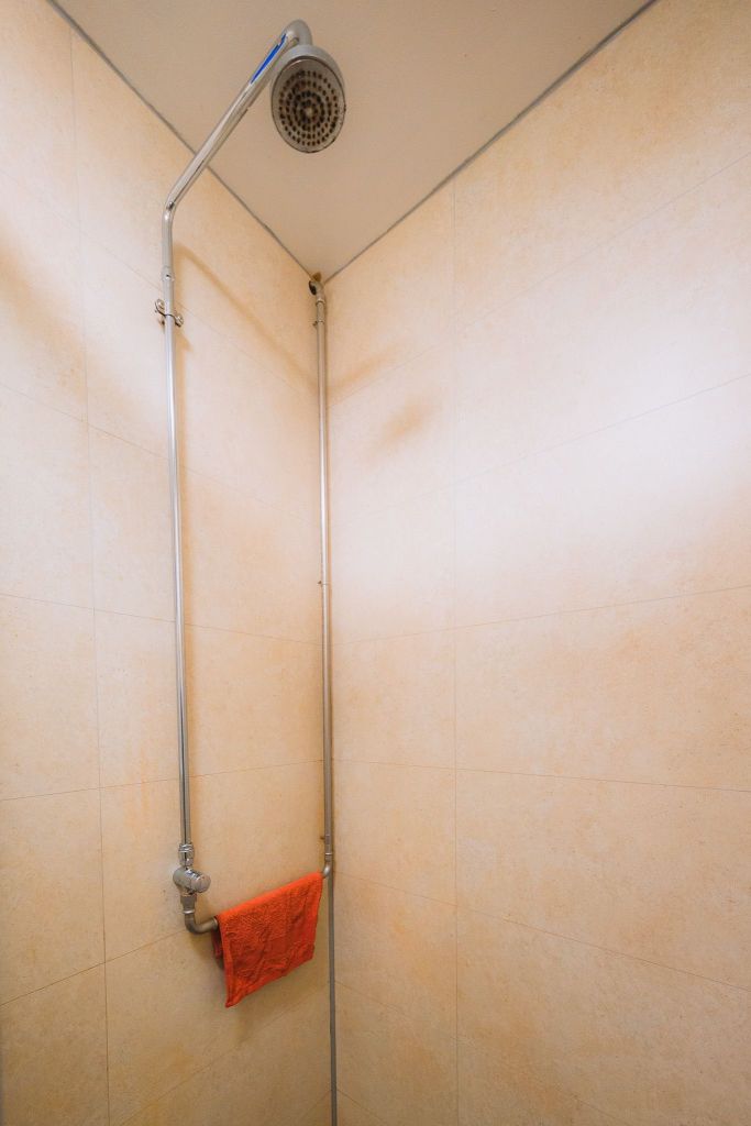 Corner of a shower with light beige tiled walls and a metal showerhead. A bright orange towel hangs on a pipe.