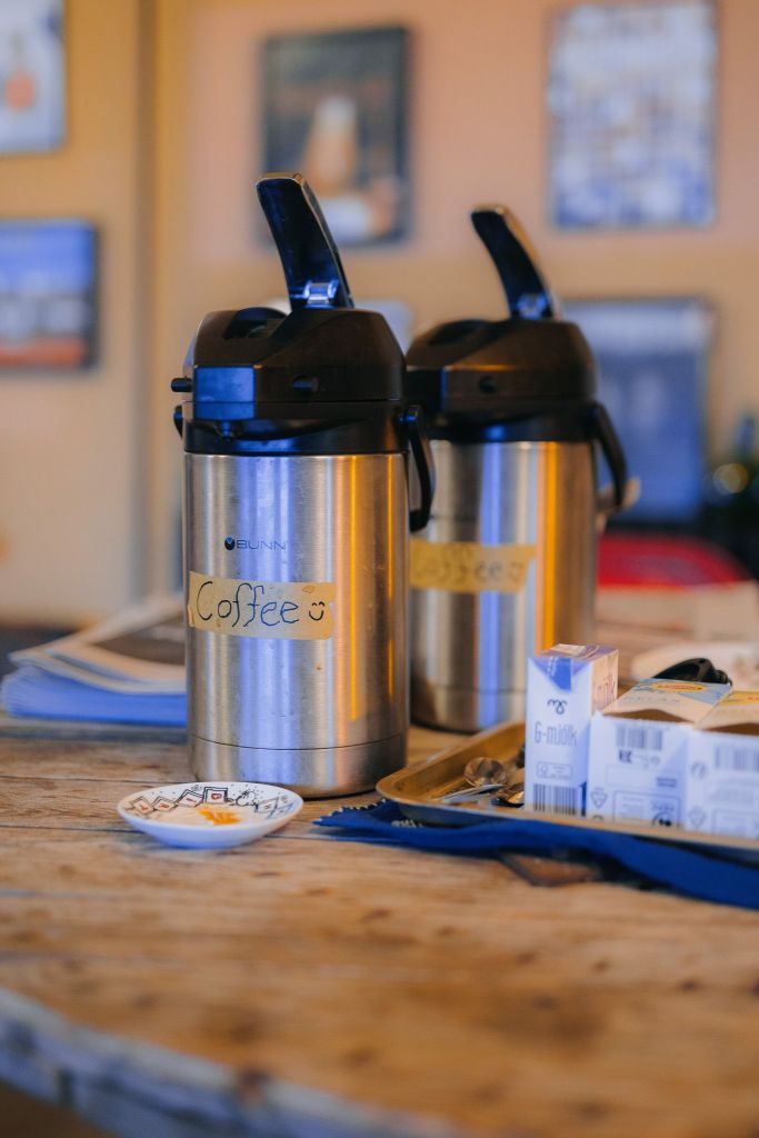 Two silver coffee airpots labeled "Coffee" sit on a rustic wooden table with milk cartons and a small dish nearby.
