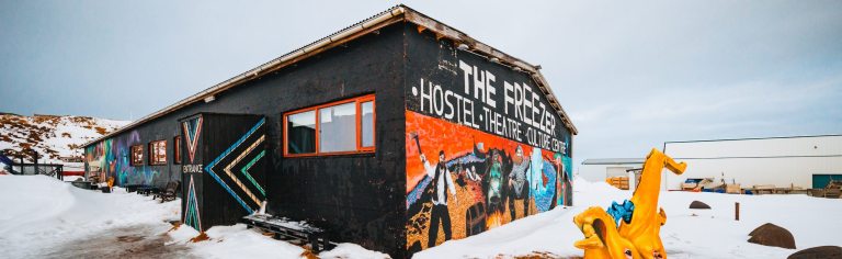 Black building in snowy landscape, featuring colorful mural with people and text "The Freezer Hostel Theatre Culture".