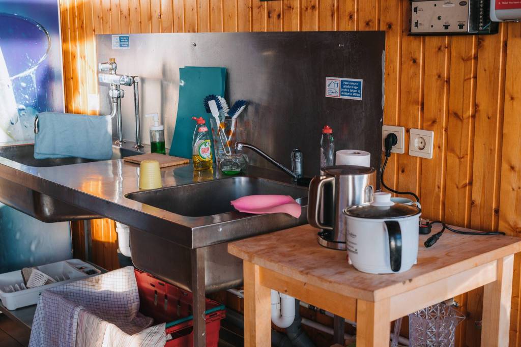 A tidy kitchen corner with a stainless steel sink, dish soap, brushes, and a drying towel. A wooden table holds a kettle and a rice cooker.
