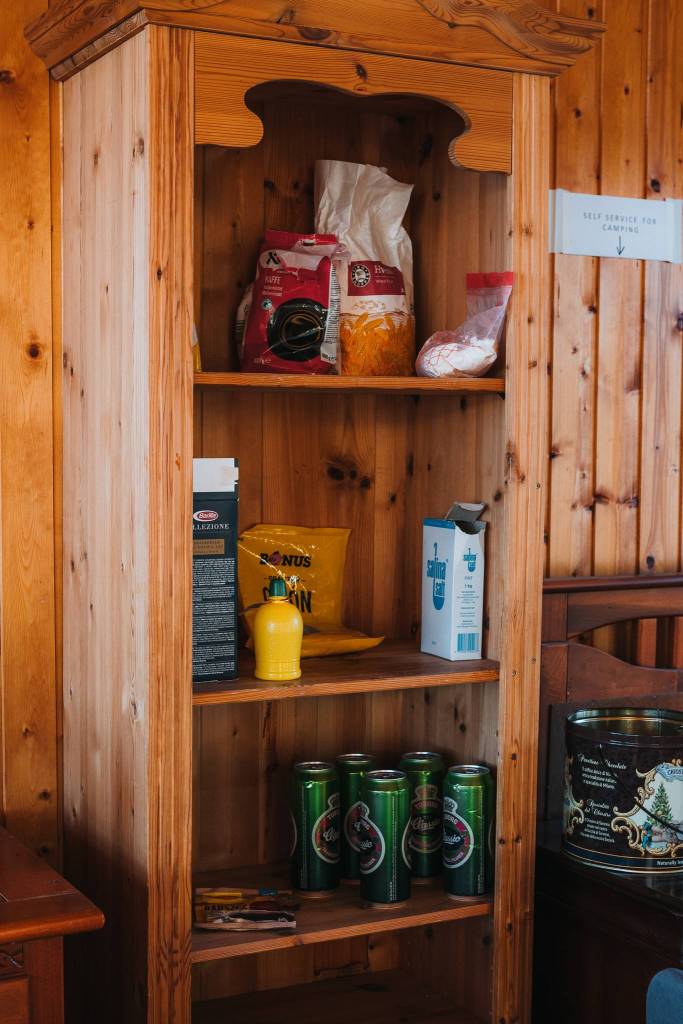 Wooden shelf with pantry items, including pasta, chips, and five green soda cans. Warm, rustic setting with a note on the wall for camping supplies.