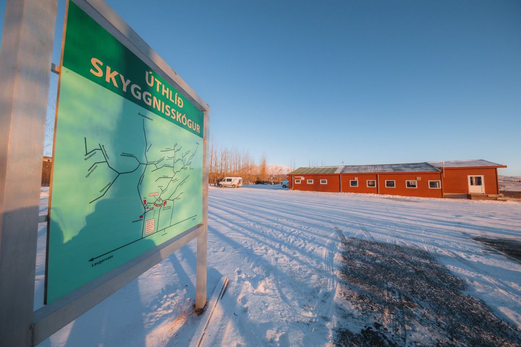 A snow-covered scene with a green map sign labeled "Úthlíð Skyggnisskógur" in the foreground. In the background, a red building stands under a clear blue sky.