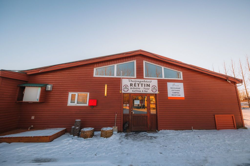 Red wooden building with large central windows and snowy ground. Sign reads "Veitingahúsið Réttin".