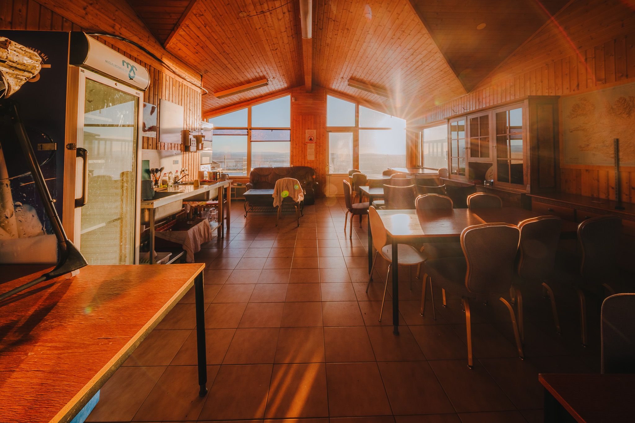 Warmly lit wooden interior of a cozy dining area with large windows, tables, and chairs.