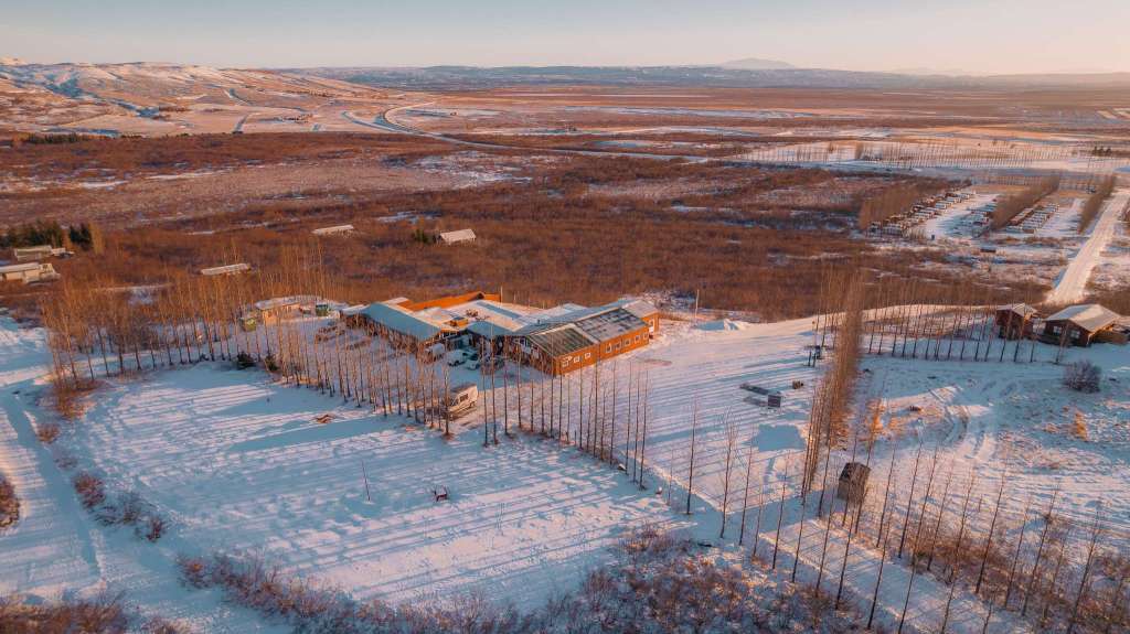 Aerial view of a snow-covered rural landscape at sunset with a cluster of buildings surrounded by trees. Rolling hills and roads stretch into the distance.