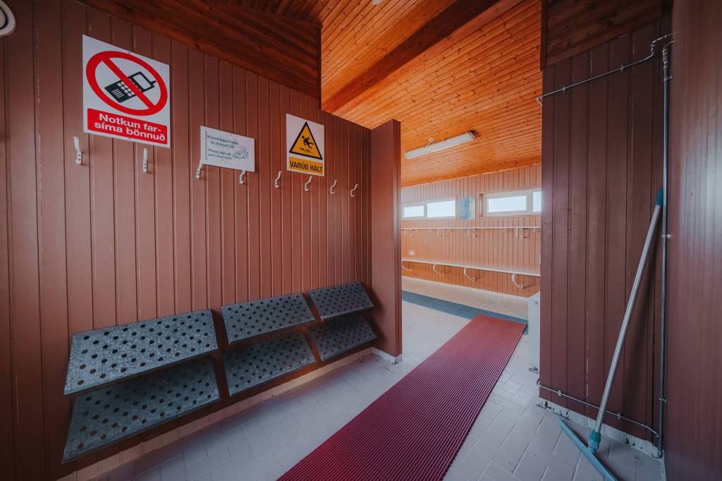 Warm-toned locker room with wooden walls, red mat on the floor, hooks on panels, and warning signs. A broom leans against the wall.