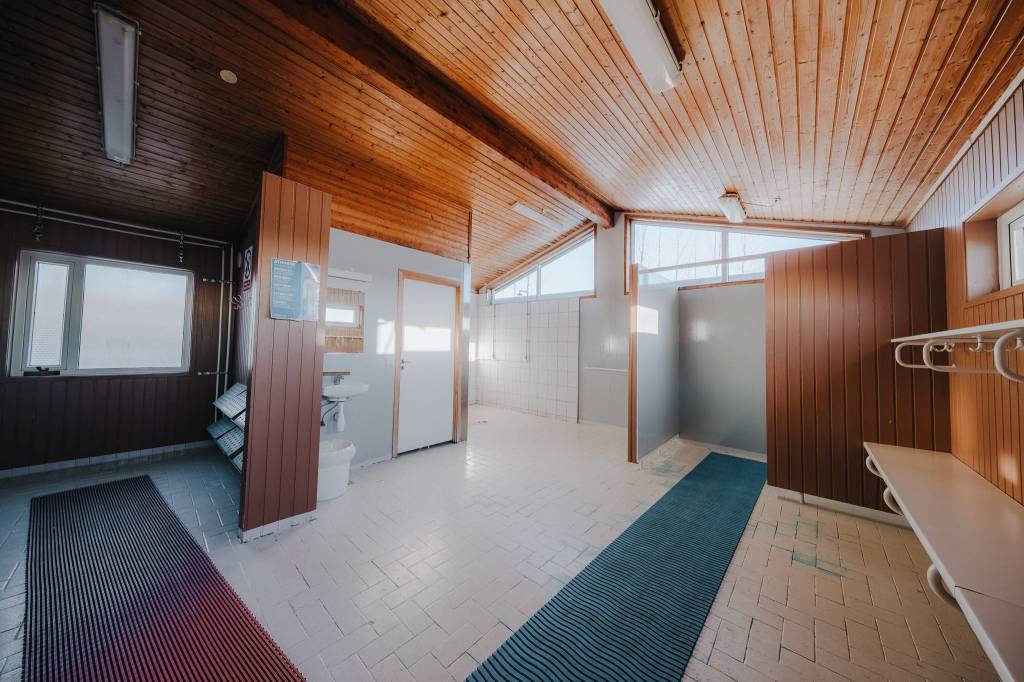 Spacious changing room with wooden ceiling and walls, white tiled floor, bench, hooks, and windows.