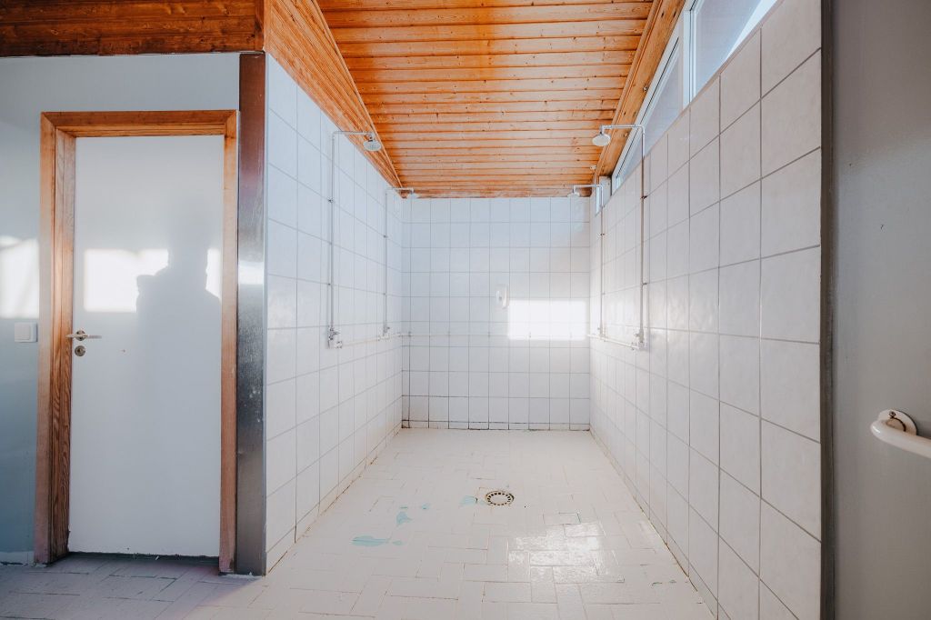 Simple, empty tiled room with a wooden ceiling and white door. Sunlight streams through a window.