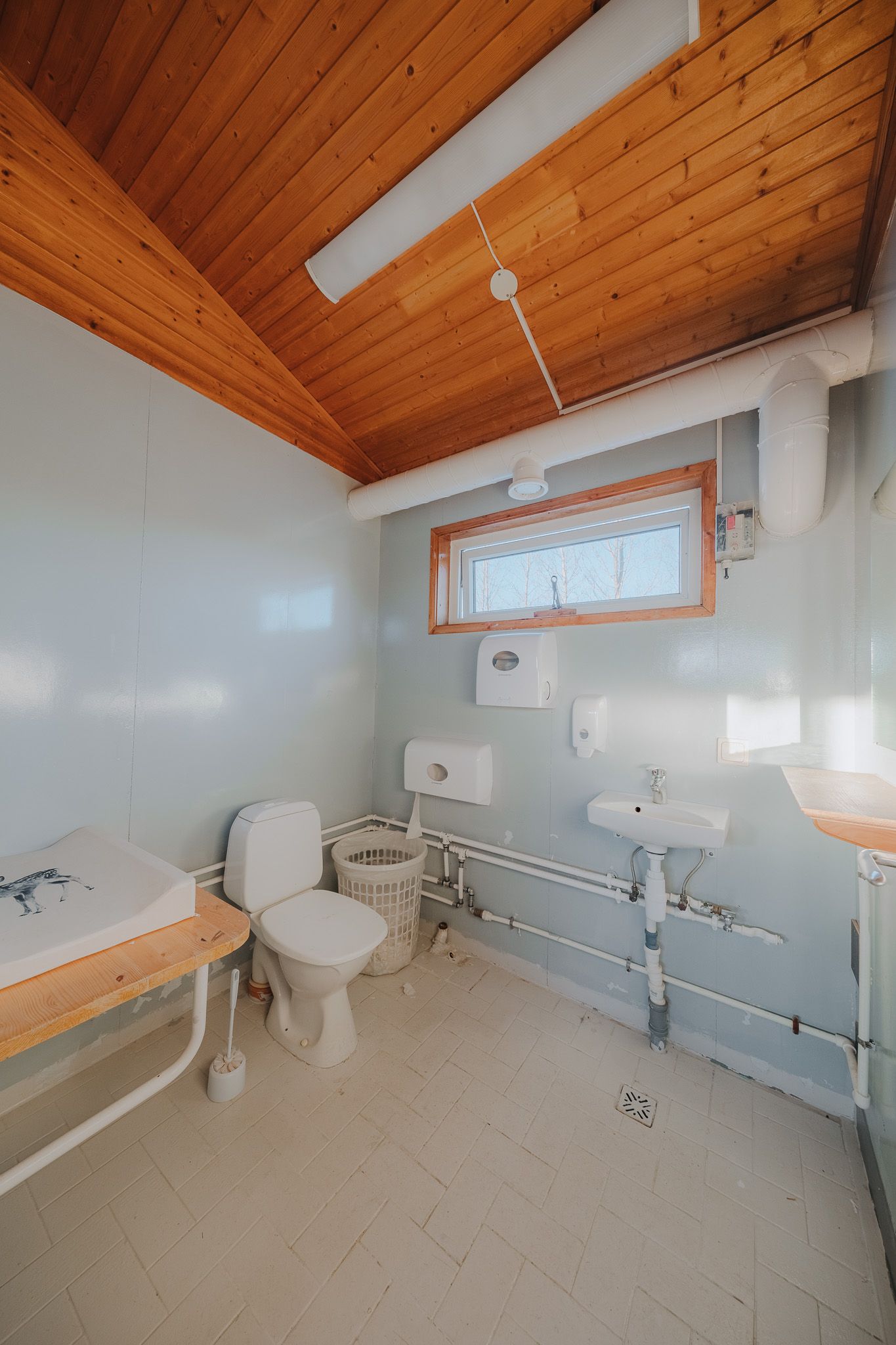 Simple restroom with wooden ceiling, toilet, changing table, and small sink.