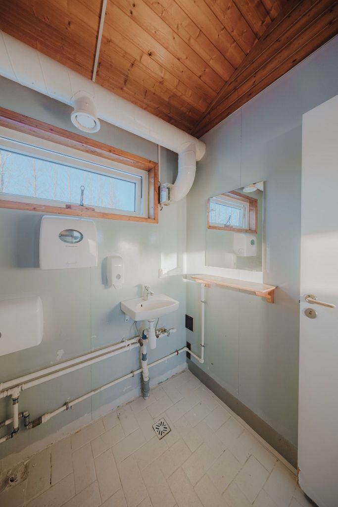 Small bathroom with light wooden ceiling and white walls. Features a small sink, hand dryer, and window.